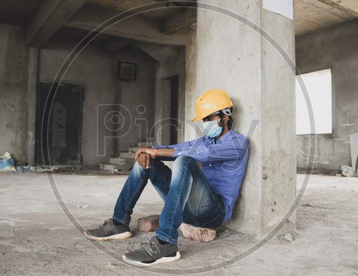 Construction Worker Sitting Sad And Lonely At Job Site Wearing A Medical Mask With Hardhat To Prevent Covid-19 Spreading, Concept Of Unemployment, Economic Crisis Job Loss During Coronavirus Crisis.