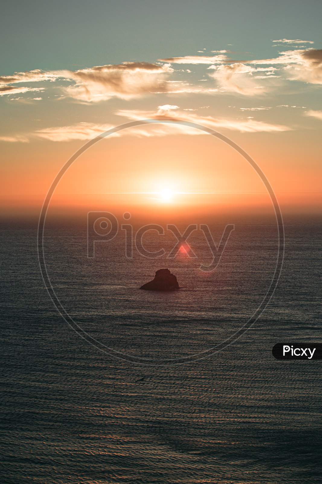 A Massive Sunset With Sun Flares Over The Ocean With A Rock In The Middle Of The Sea