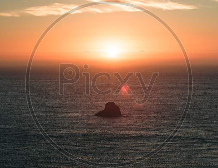 A Massive Sunset With Sun Flares Over The Ocean With A Rock In The Middle Of The Sea