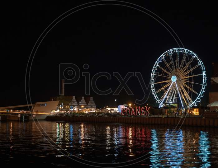 Gdansk, North Poland - August 13, 2020: Night Photography Of Cityscape Consisting Old Town Commercial Market And Illuminated Ferries Wheel Over Motlawa River