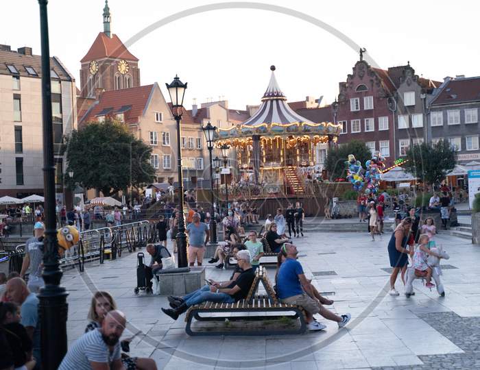Gdansk, North Poland - August 13, 2020: People Doing Leisure At City Center Opposite Carousel Horses At The Old Town Over Motlawa River