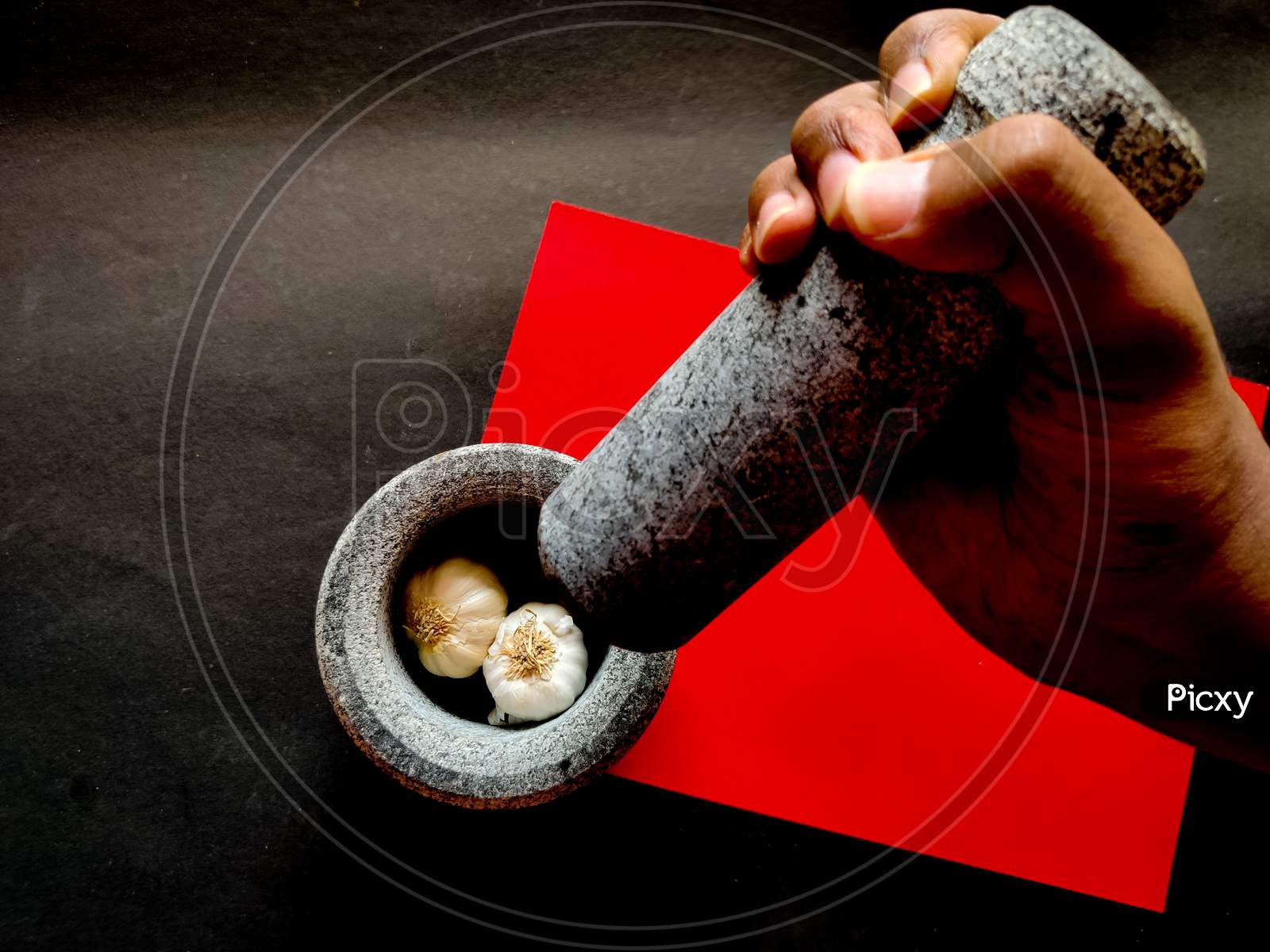 Human Hand Holding Mortar And Ready To Crush The Garlic In Pestle. Isolated On Red And Black Background.