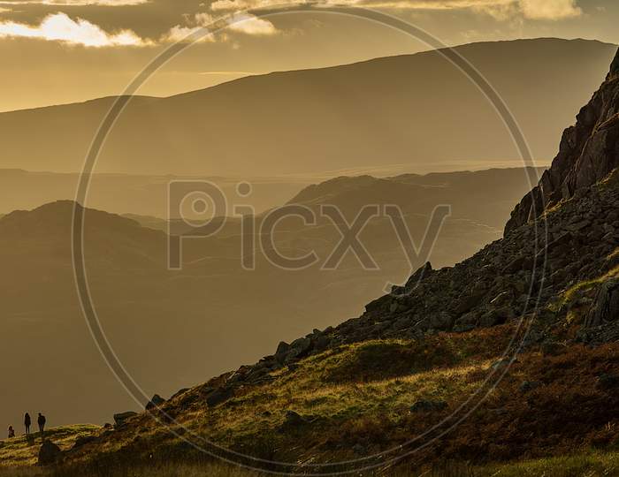 A Group Of Walkers Are Silhouetted Against A Hazy Yellow Backdrop Of Lakeland Fells