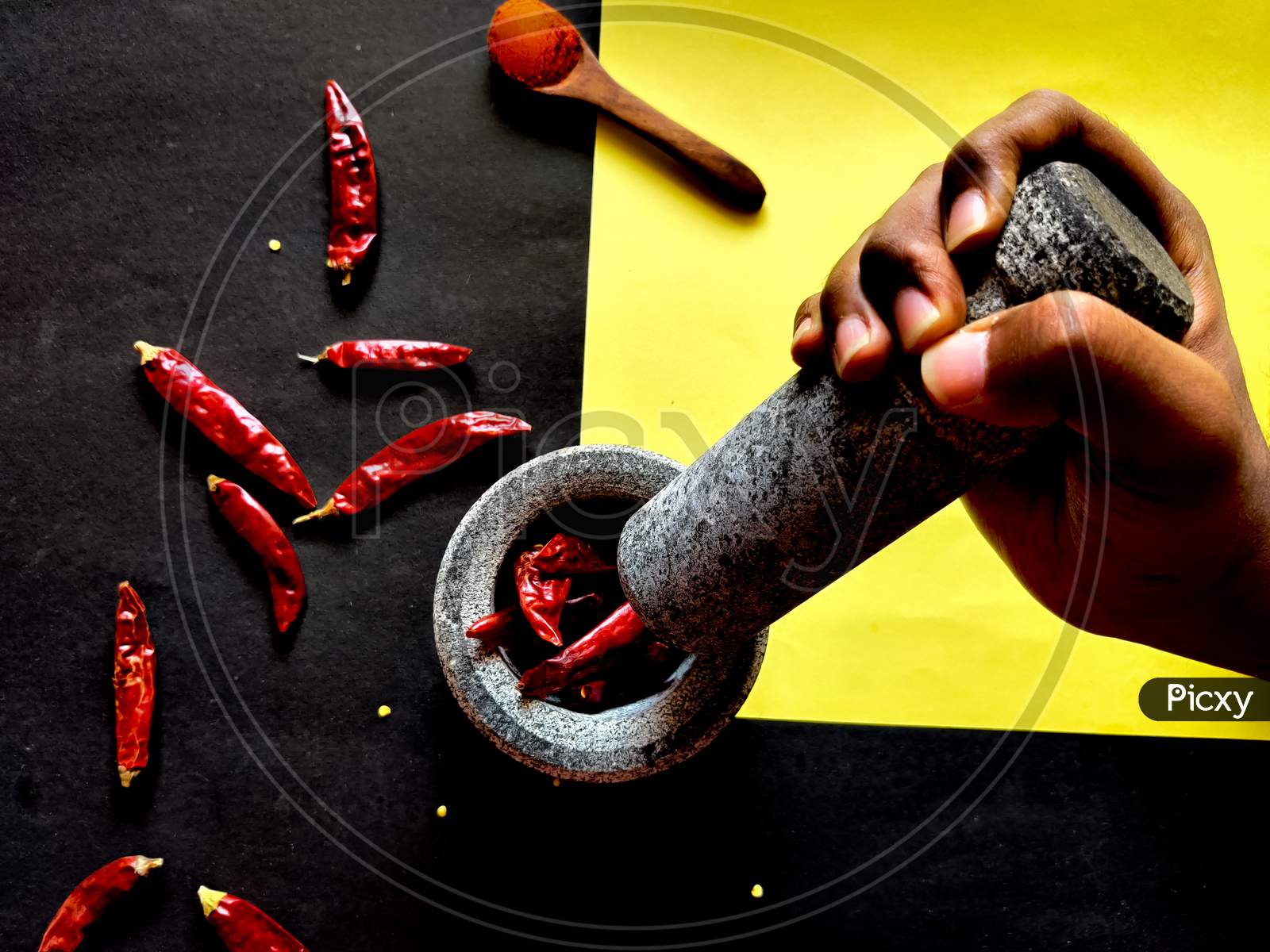 Human Hand Holding Mortar And Ready To Crush The Red Chillies In Pestle. Isolated On Yellow And Black Background.