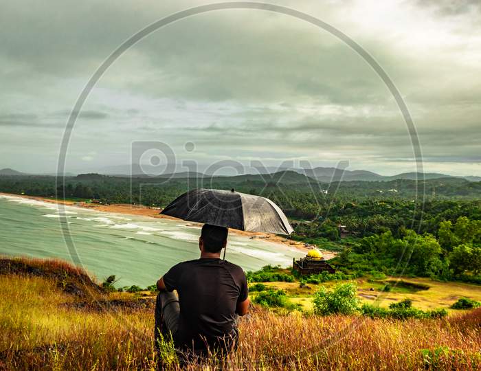 Man Sitting On Hilltop With Umbrella Enjoying Rain With Amazing View