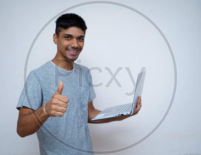 Young Indian Handsome Boy Showing Thumbs Up And Smiling While Looking Into The Camera, Holding Laptop In His Other Hand.