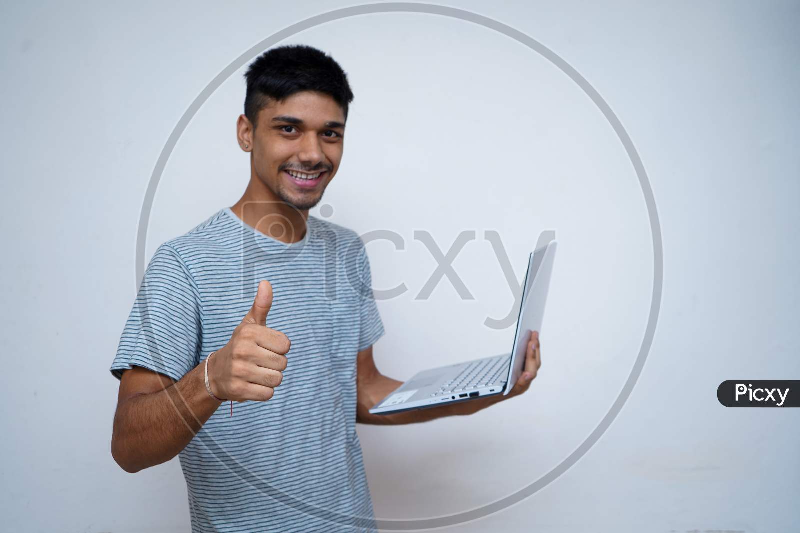 Young Indian Handsome Boy Showing Thumbs Up And Smiling While Looking Into The Camera, Holding Laptop In His Other Hand.