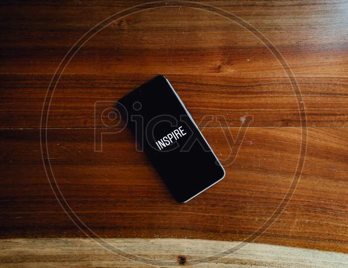 Flat Lay Of A Black Phone Over A Wood Background With The Word Inspire Written In It