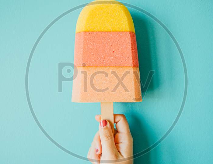Super Minimalistic Hand Grabbing A Colorful Foam Ice Cream In Front Of A Pastel Blue Wall