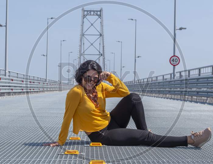 Smiling Woman Sitting In The Middle Of The Street Take Off Sunglasses While Looking At The Camera Over Bridge Background.