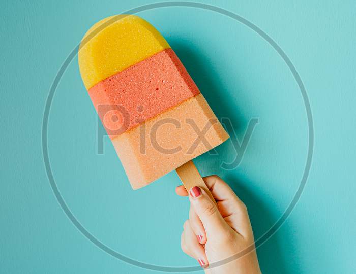 Super Minimalistic Hand Grabbing A Colorful Foam Ice Cream In Front Of A Pastel Blue Wall