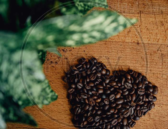 Flat Lay Of A Heart Symbol Made Of Coffee Grains Over A Wood Table With Some Leaves Decorating