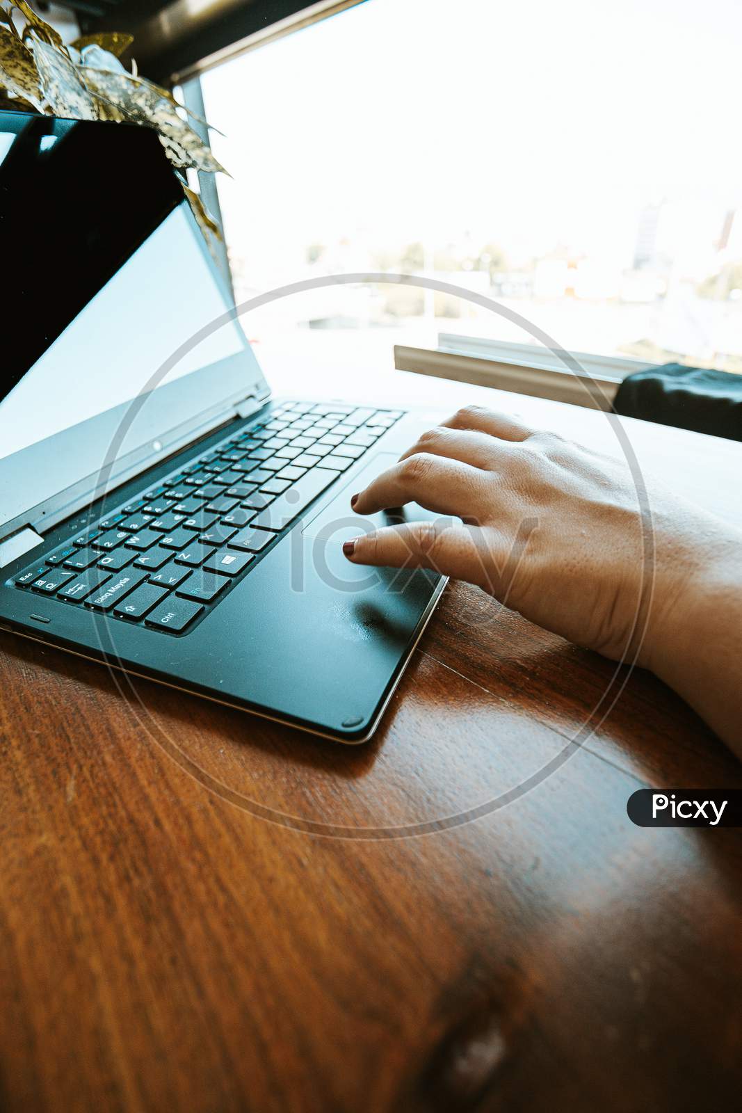 Female Hand Working On A Black Laptop Over A Wooden Table During A Bright Day