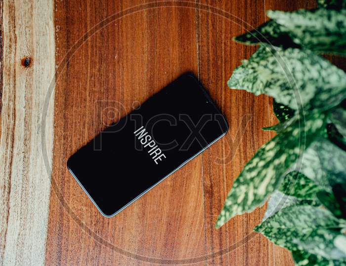 Flat Lay Of A Black Phone Over A Wood Background And Some Leaves Near It With The Word Inspire Written In It