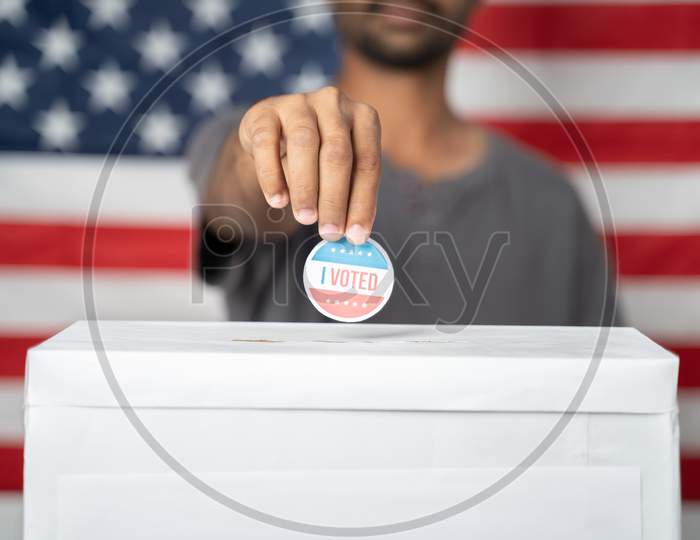 Concept Of Usa Elections, Close Up Of Hands Putting I Voted Sticker Inside Ballot Box With Us Flag As Background.