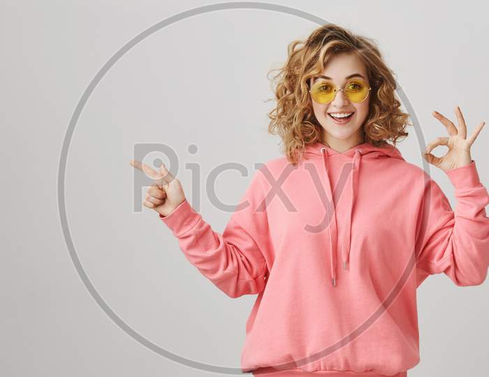 I Am Okay With That. Studio Shot Of Good-Looking Curly-Haired Girl With Bright Smile Showing Fine Gesture And Pointing Left With Friendly And Happy Expression, Standing Over Gray Background.