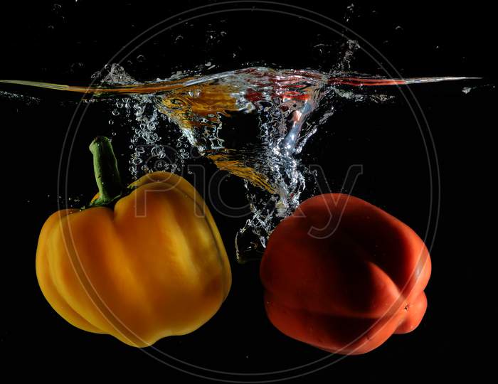 Red And Yellow Capsicum Dips Into Water Creating Water Splash With a dark Or Black Ground