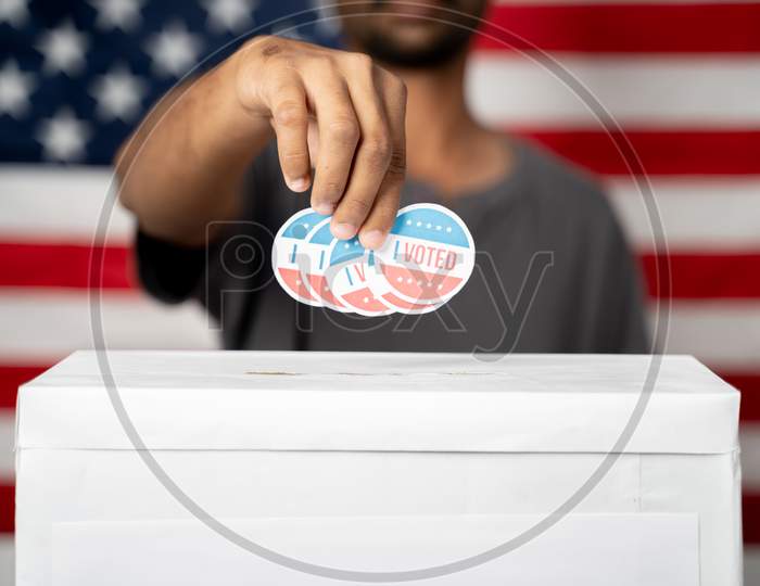 Close Up Of Hands Dropping Multiple I Voted Sticker Inside Ballot Box With Us Flag As Background, Concept Of Fraud In Usa Elections.