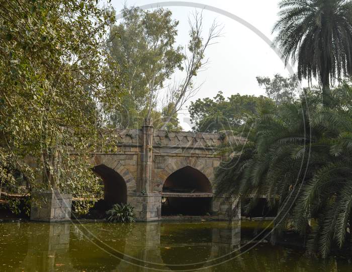 A Reflection And Mesmerizing View From The Side Of The Pond,Lake Of Palm Trees And Bridge Monument At Lodi Garden Or Lodhi Gardens In A City Park At Winter Foggy Morning.