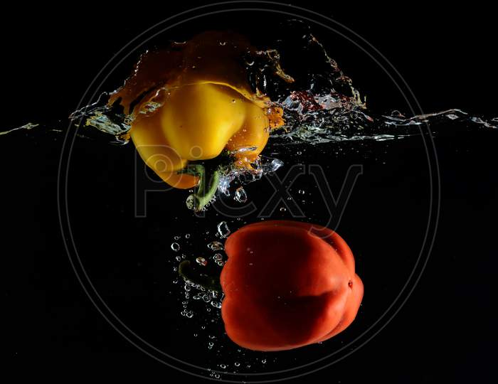 Red And Yellow Capsicum Dips Into Water Creating Water Splash With a dark Or Black Ground