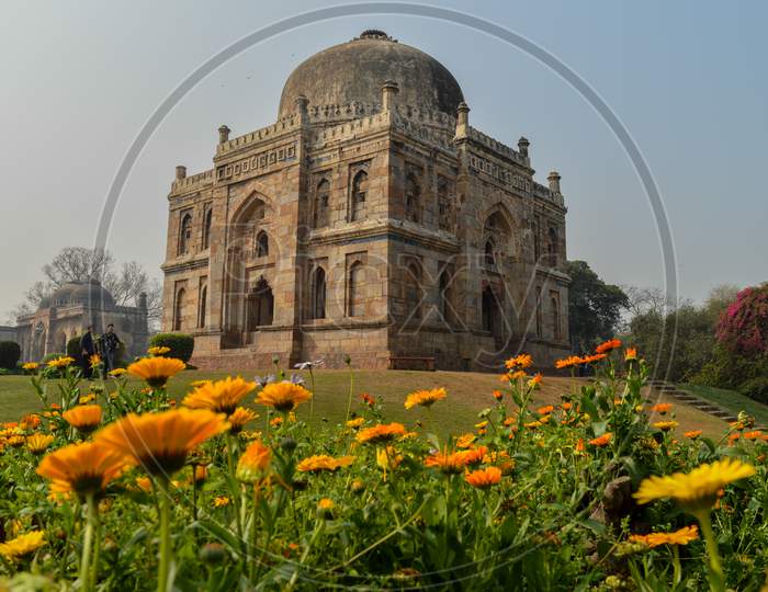 A Mesmerizing View Of Shish Gumbad Monument With Yellow Flowers At Lodi Garden Or Lodhi Gardens In A City Park From The Side Of The Lawn At Winter Foggy Morning.