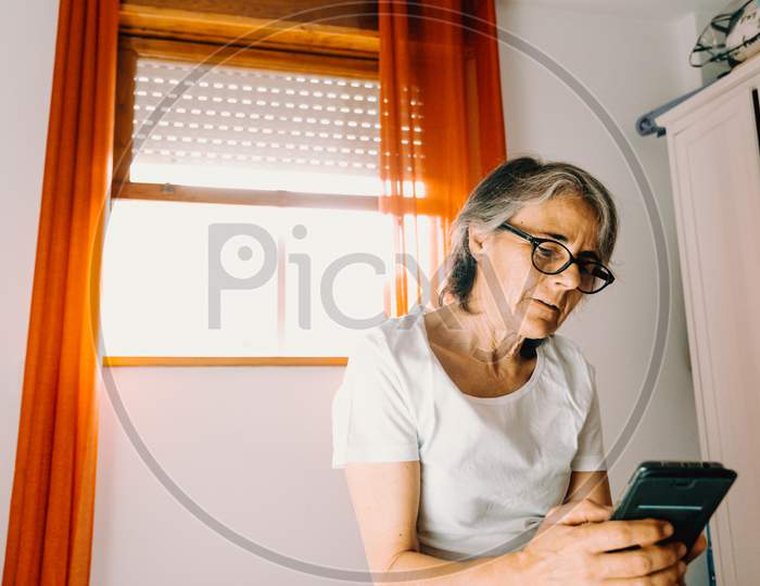 Old Woman With Glasses Checking His Phone And Paying Attention In His Bedroom