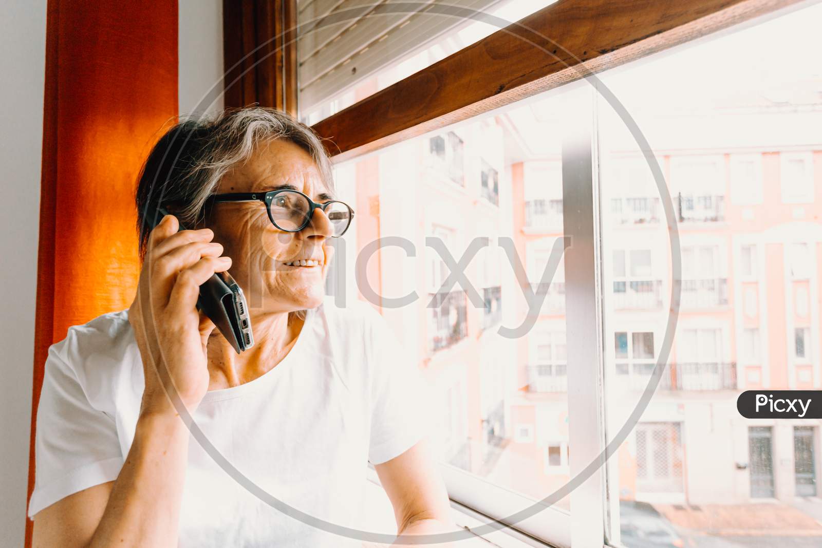 Close Up Of An Old Woman Making A Call On The Mobile Phone And Smiling While Looking Through The Window