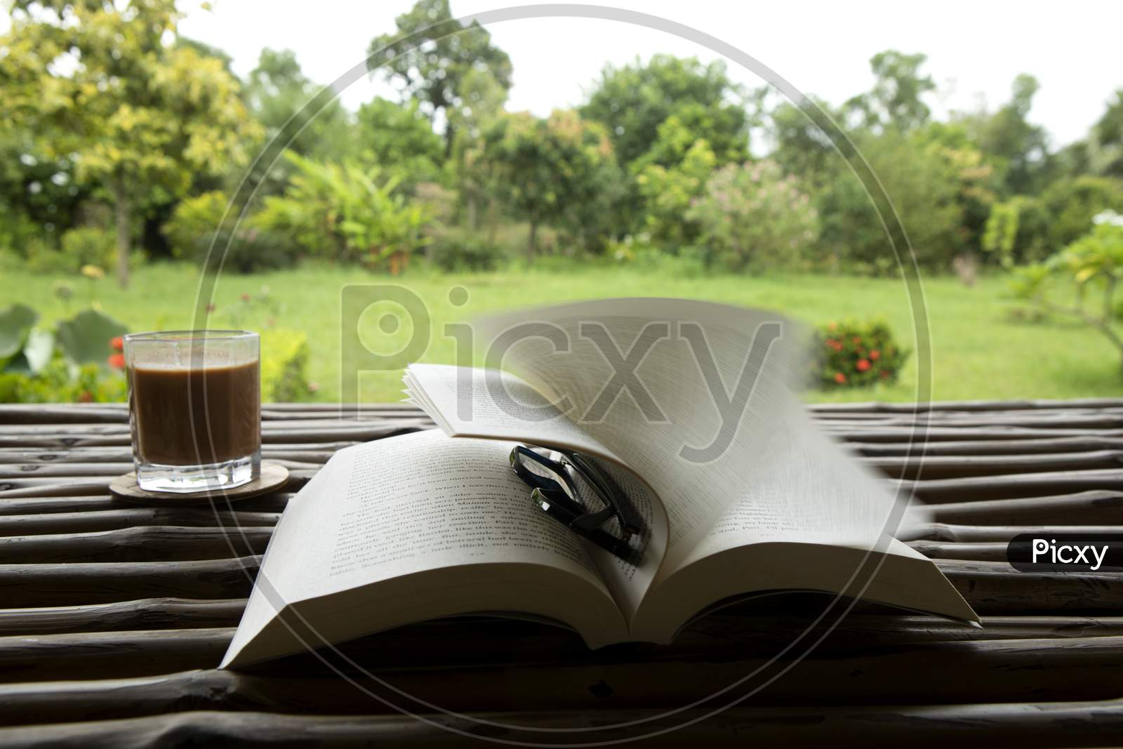 Tea In A Glass With A Book And A Spectacle On A Bamboo Table In A Beautiful Green Garden In A Windy Monsoon Morning