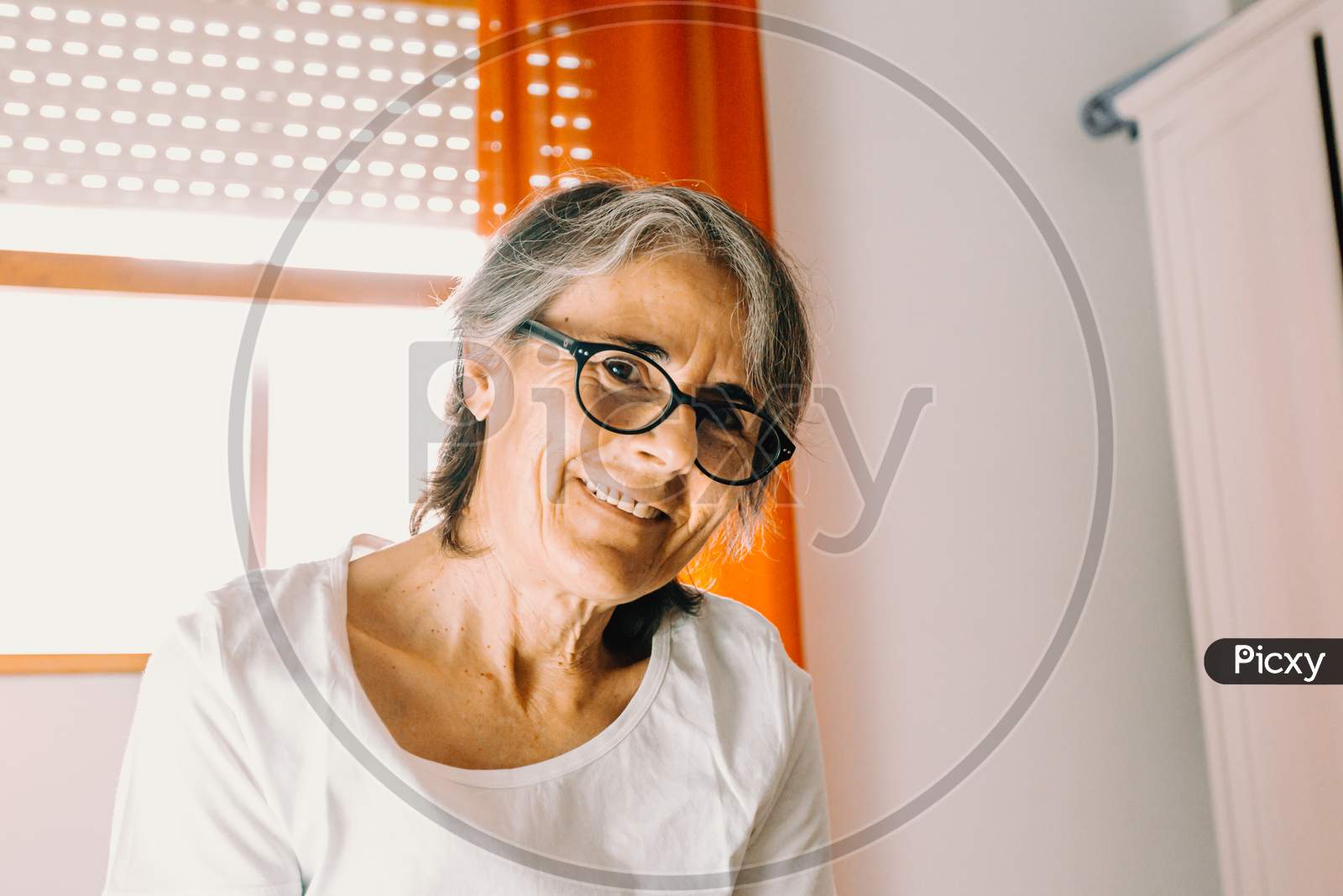 Close Up Of A Old Woman With Glasses Smiling To Camera In A Bright Bedroom
