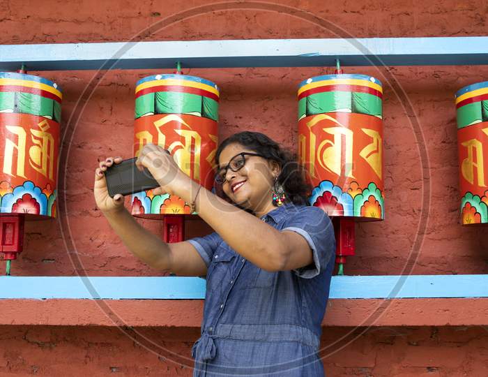 A Beautiful Lady In A Blue Dress Taking Selfie With A Smart Phone In Front Of Buddhist Prayer Wheels In A Monastery In Sikkim In India