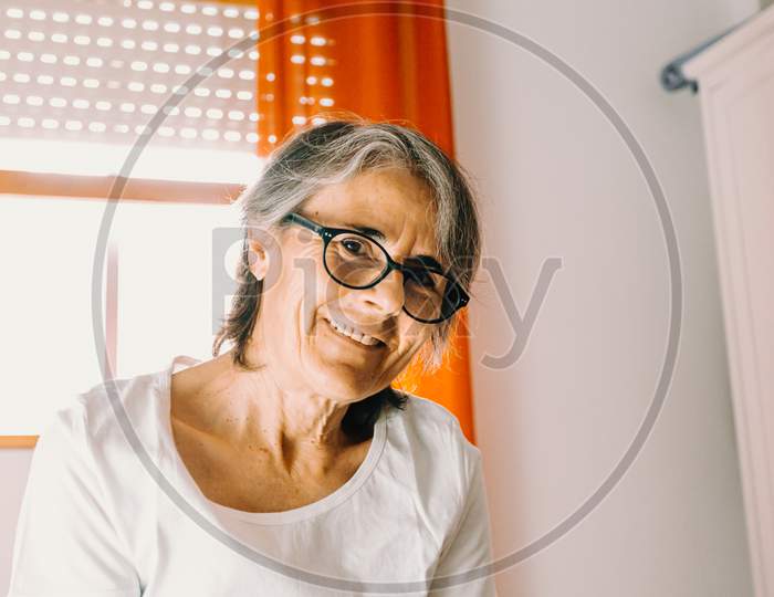 Close Up Of A Old Woman With Glasses Smiling To Camera In A Bright Bedroom