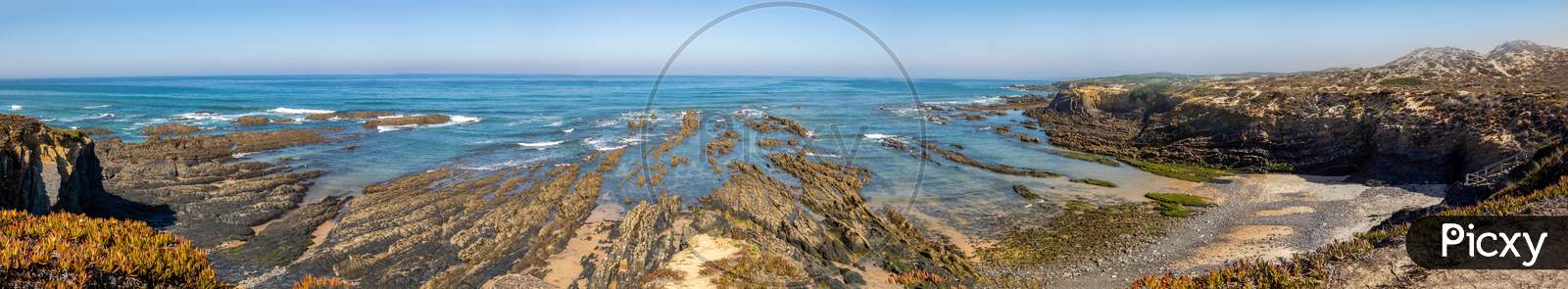 Wide panoramic view of beach in Costa Vicentina, Alentejo. Rocky beach, Atlantic ocean, cliff side with rocks. Amazing blue water and clear blue sky.