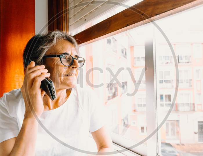 Close Up Of An Old Woman Making A Call On The Mobile Phone And Smiling While Looking Through The Window