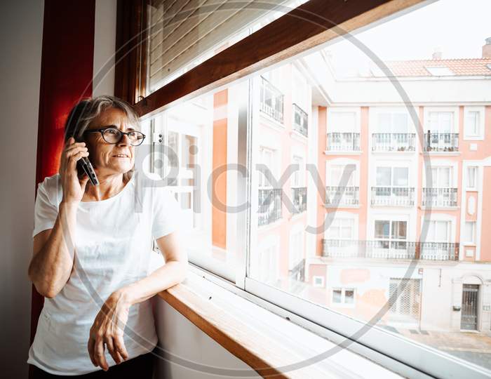Old Woman Making A Call On The Mobile Phone While Looking Through The Window