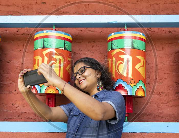 A Beautiful Lady In A Blue Dress Taking Selfie With A Smart Phone In Front Of Buddhist Prayer Wheels In A Monastery In Sikkim In India