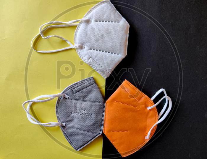 Three Kn-95 Face Masks In White,Grey And Orange Color. Isolated On Black And Yellow Background. Protection From Coronavirus Or Covid-19. Wear A Mask. Coronavirus Pandemic. Top View
