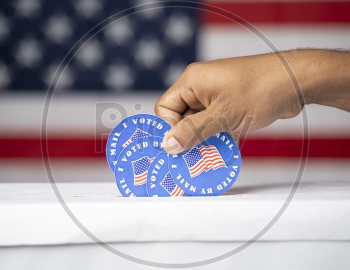Close Up Of Hands Putting Multiple I Voted My Mail Stickers Inside The Box With Us Flag As Background - Concept Of Vote By Mail Fraud In Usa Elections.