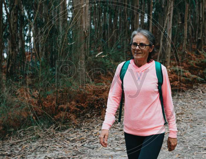 Close Up Of An Old Woman Walking In The Forest With Sport Clothes, Bag And Glasses While Smiling