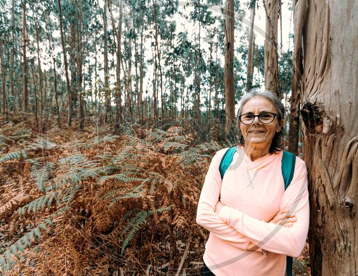 Old Woman Leaning On A Tree While Smiling To Camera In The Forest On Sport Clothes