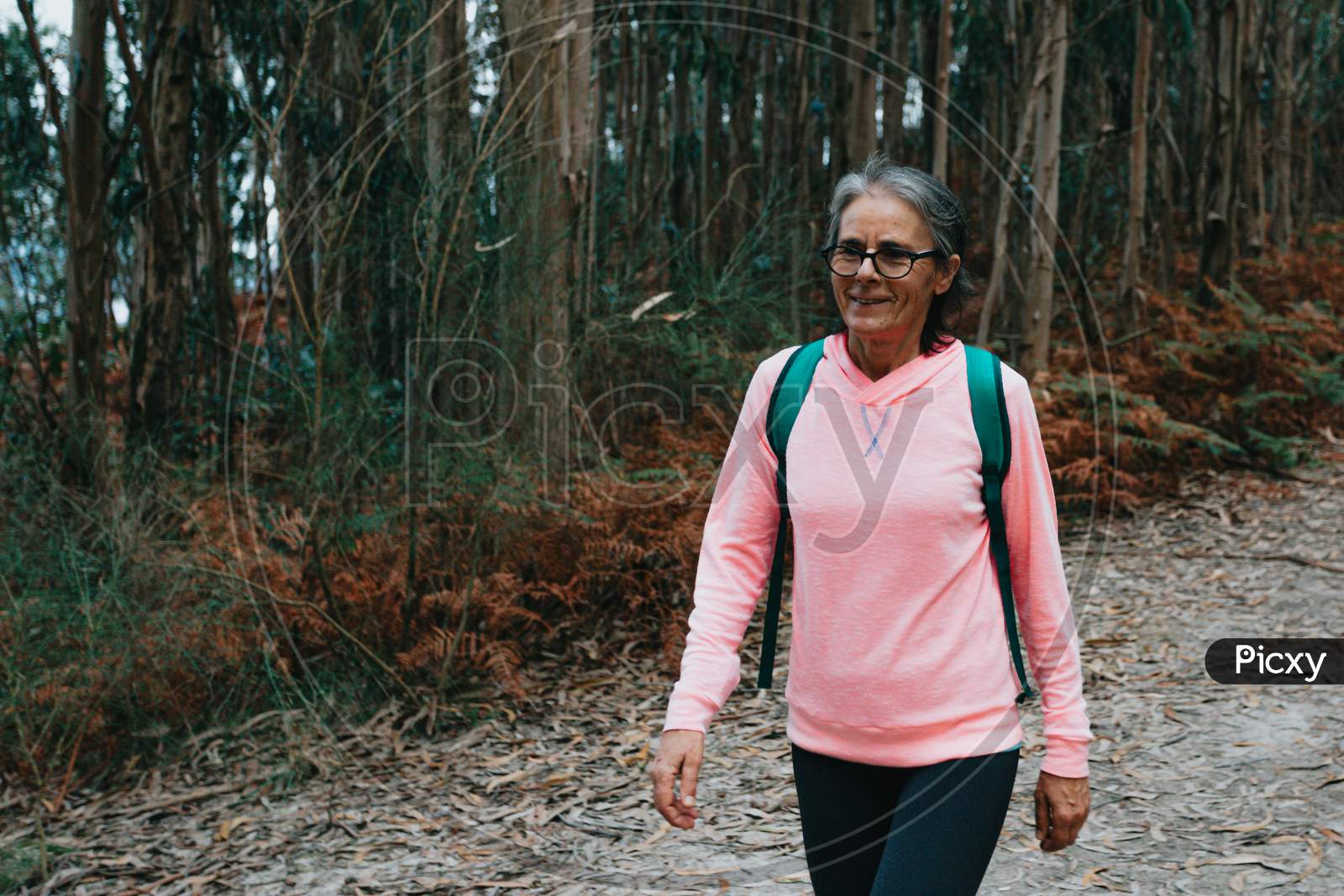 Close Up Of An Old Woman Walking In The Forest With Sport Clothes, Bag And Glasses While Smiling