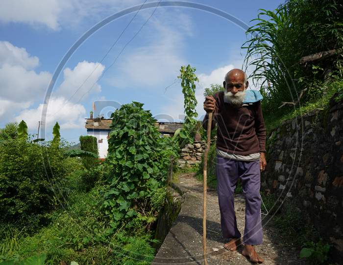 An Old Man With Long White Beard Walking Down A Narrow Pathway With The Help Of A Stick.