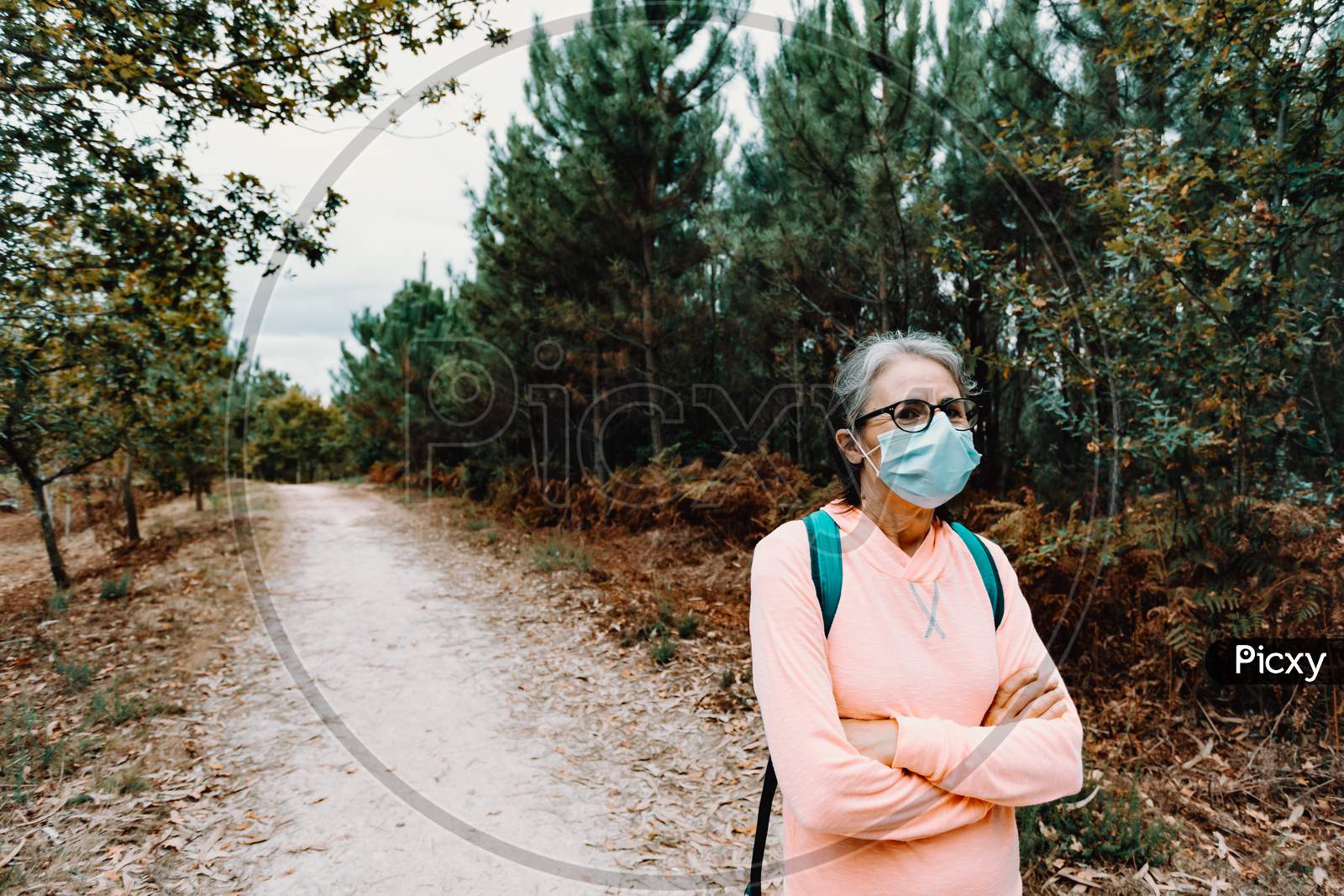 Old Woman Crossing Arms While Wearing A Mask In The Middle Of A Forest With Copy Space