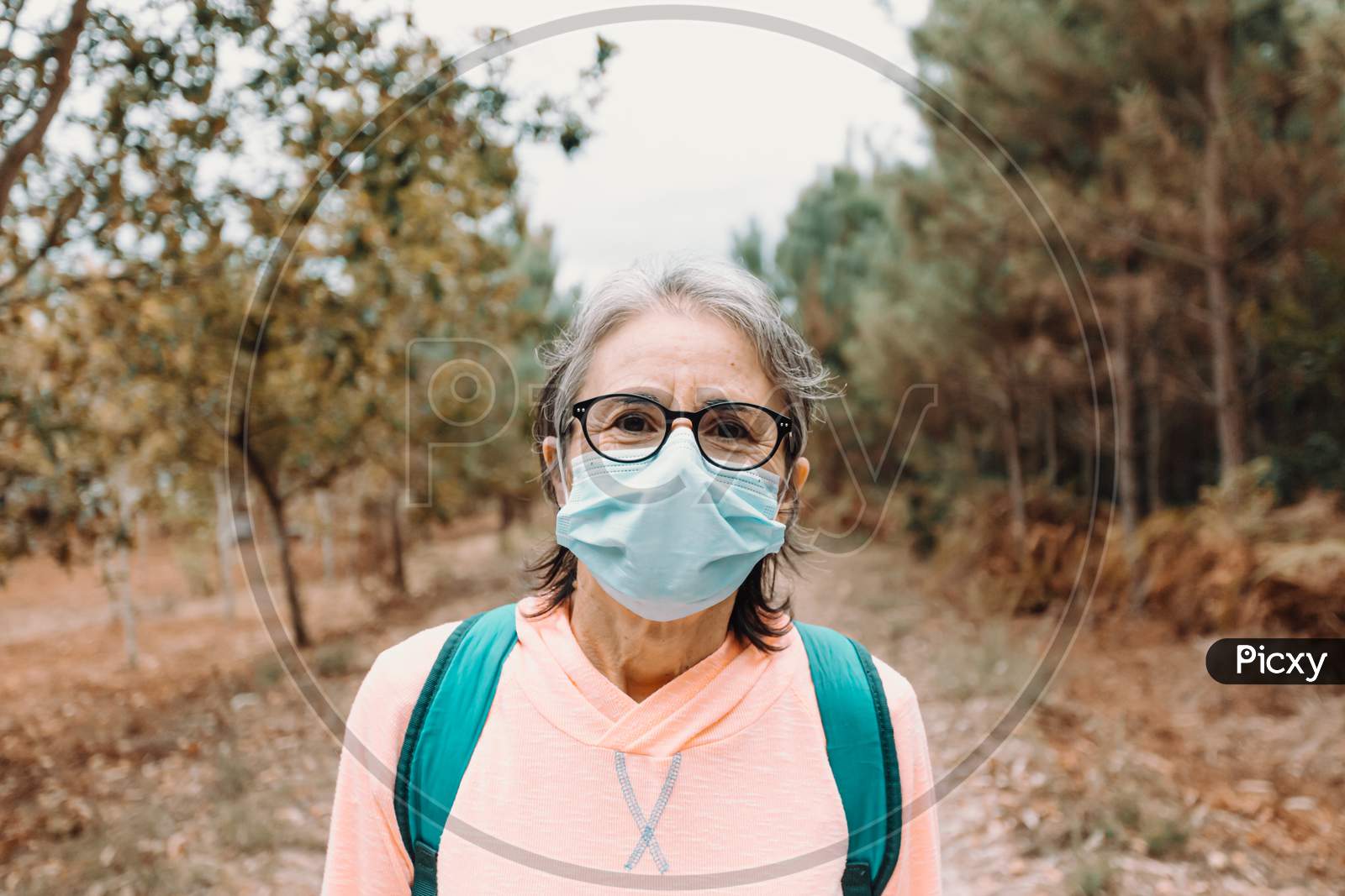 Close Up Of A Frontal Portrait Of An Old Woman Wearing A Mask And Sport Clothes In The Forest During A Trekking Day