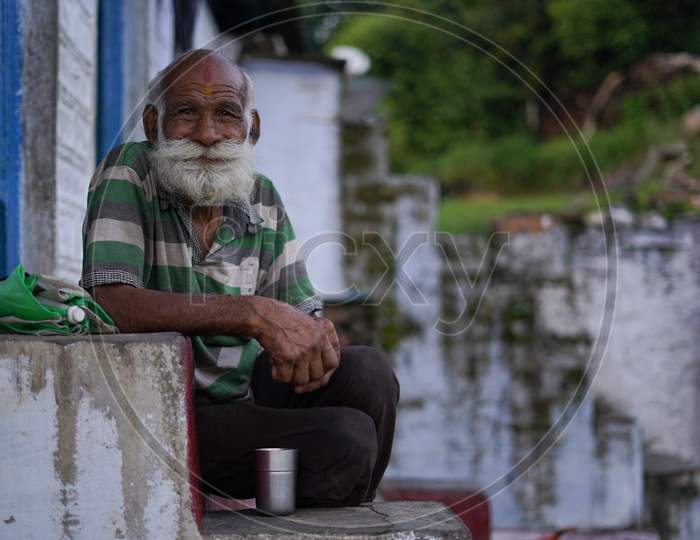 An Old Indian Man With Long And White Beard Sitting On The Stairs Wearing Rugged Clothes.