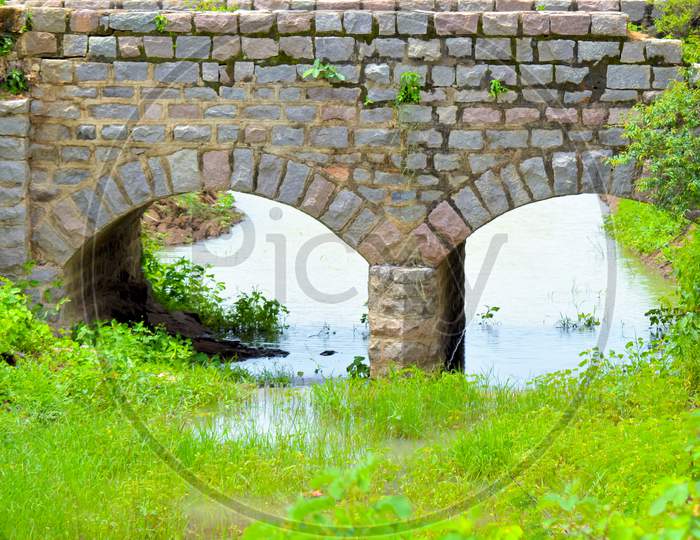 Old Stone Single Arch Bridge, Latvia. Famous Ancient Stone Arch Single Track Road Bridge In The Forest. View Of Small River And Trees Reflection In The Water.