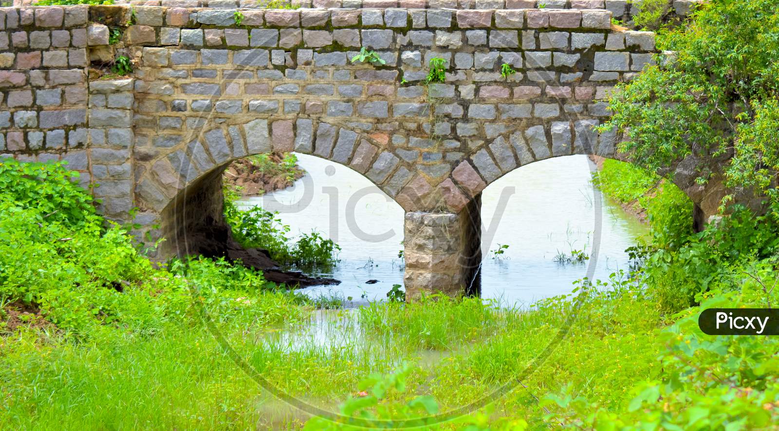 Old Stone Single Arch Bridge, Latvia. Famous Ancient Stone Arch Single Track Road Bridge In The Forest. View Of Small River And Trees Reflection In The Water.