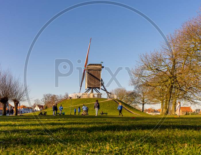 Belgium, Bruges, A Group Of People Standing On Top Of A Grass Covered Field Near Windmill