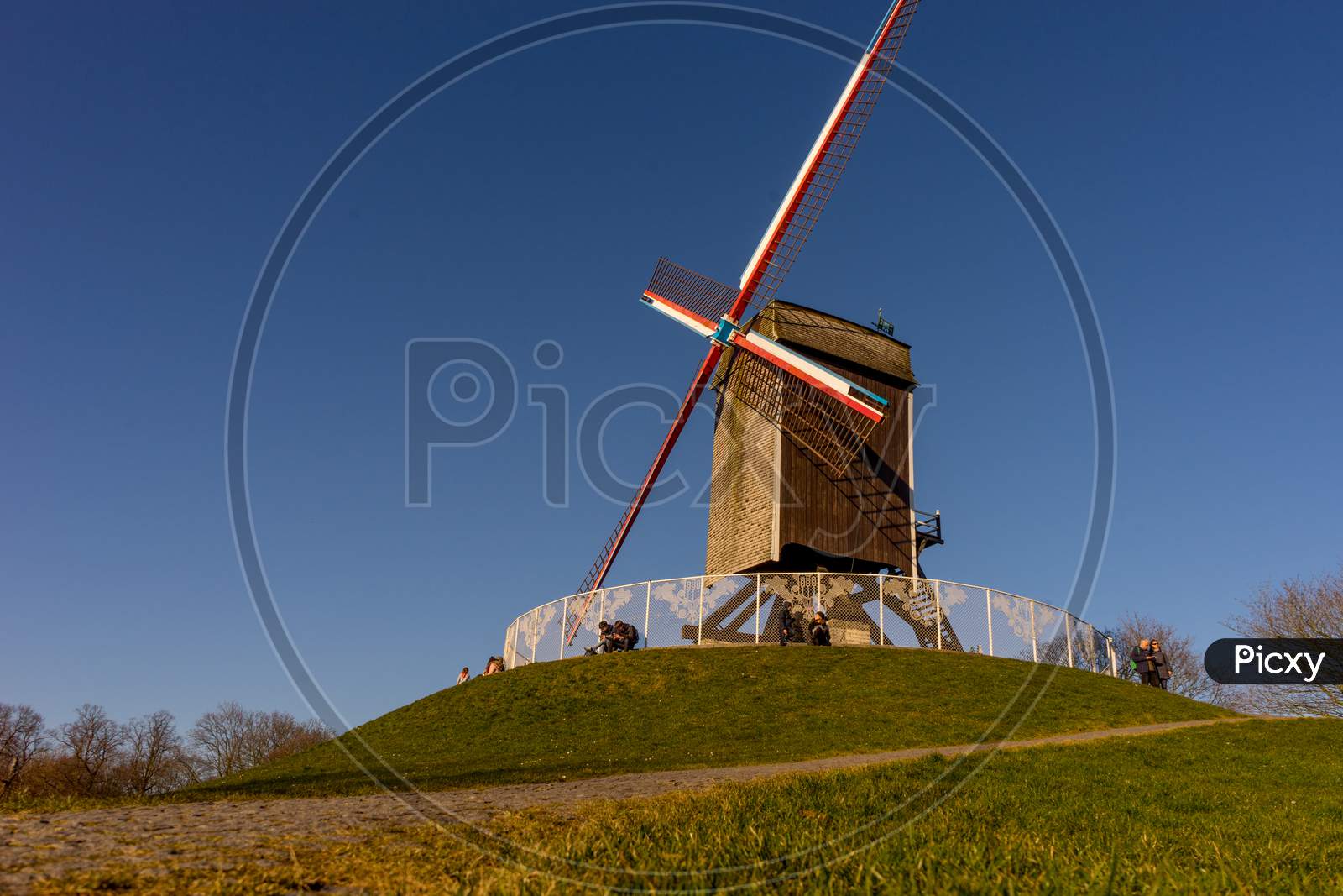 Belgium, Bruges, A Clock Tower On Top Of A Grass Covered Field With Windmill Island In The Background