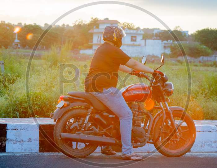 Young Indian Man With Full Face Helmet And Protective Gloves Riding A Red Royal Enfield At Sunset Golden Hour