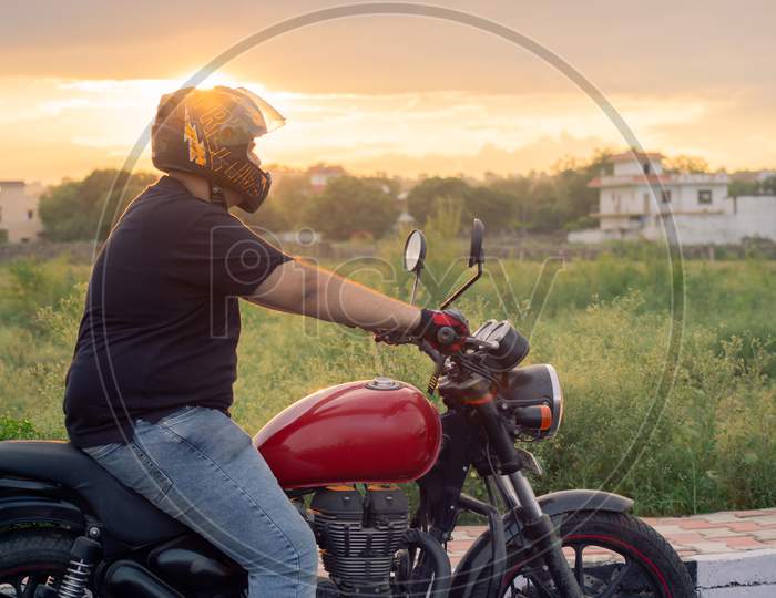 Young Indian Man With Full Face Helmet And Protective Gloves Riding A Red Royal Enfield At Sunset Golden Hour